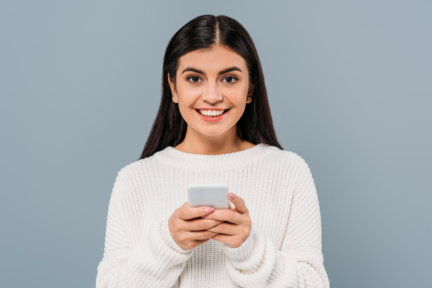 glimlachen mooi brunette meisje in wit trui met behulp van smartphone geïsoleerd op grijs - Foto, afbeelding