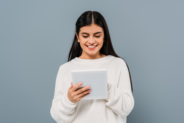 uśmiechnięta ładna brunetka dziewczyna w białym swetrze za pomocą cyfrowej tabletki izolowane na szarości - Zdjęcie, obraz
