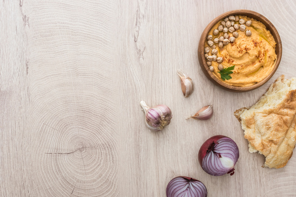 Blick von oben auf köstlichen Hummus mit Kichererbsen in Schüssel in der Nähe von frisch gebackener Pita, Knoblauch und roter Zwiebel auf beigem Holztisch - Foto, Bild