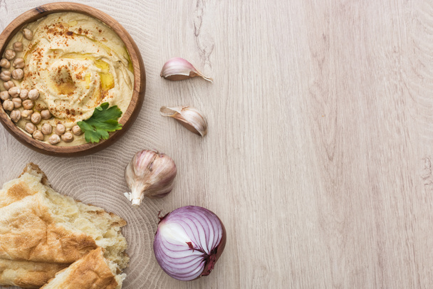 vista superior del delicioso hummus con garbanzos en un tazón cerca de pita recién horneada, ajo y cebolla roja en una mesa de madera beige
 - Foto, imagen
