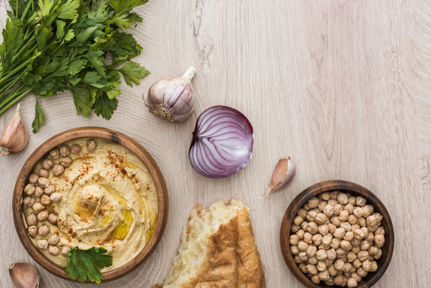 top view of delicious hummus with chickpeas in bowl near fresh baked pita, garlic, parsley and red onion on beige wooden table - Фото, изображение