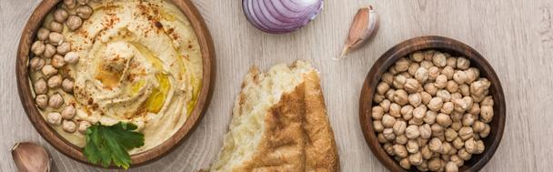 bovenaanzicht van heerlijke hummus met kikkererwten in kom bij vers gebakken pita, knoflook en rode ui op beige houten tafel, panoramisch schot - Foto, afbeelding