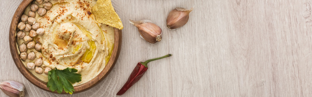 vrchní pohled na lahodný hummus s cizrnou, nacho v misce poblíž koření a petrželky na béžovém dřevěném stole, panoramatický záběr - Fotografie, Obrázek