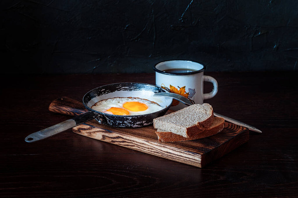Fried scrambled eggs in vintage pan, with bread, old fork, metal mug with tea, side view, dark background - Photo, Image