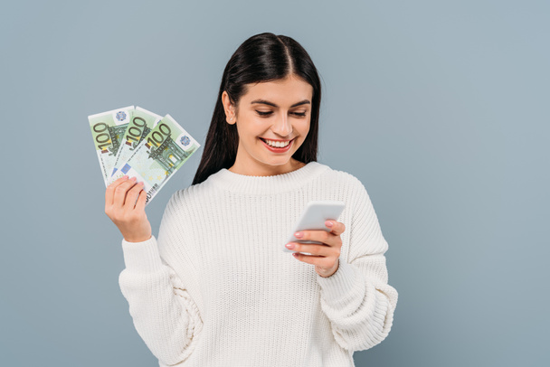 niña bonita sonriente en suéter blanco con billetes en euros y el uso de un teléfono inteligente aislado en gris
 - Foto, Imagen