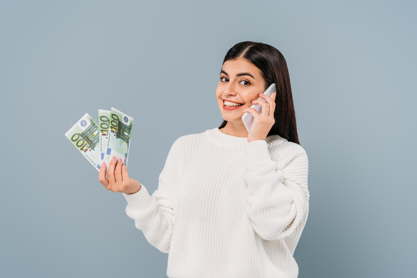 улыбающаяся красивая девушка в белом свитере, держащая банкноты евро и разговаривающая на смартфоне, изолированном на сером
 - Фото, изображение