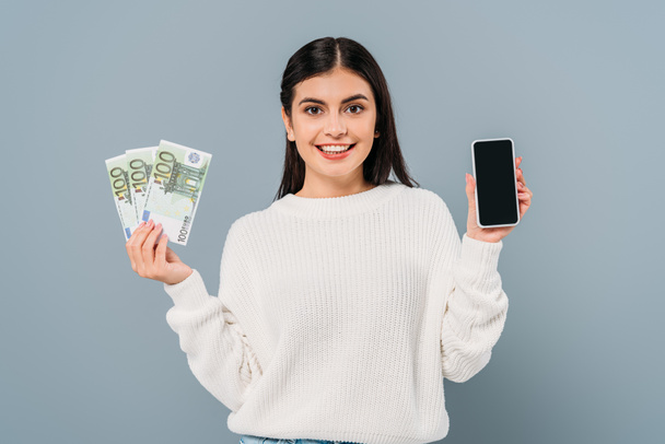 usmívající se hezká dívka v bílém svetru drží eurobankovky a smartphone s prázdnou obrazovkou izolované na šedé - Fotografie, Obrázek