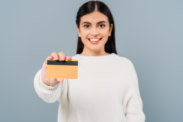 selective focus of smiling pretty girl in white sweater presenting credit card isolated on grey - Fotoğraf, Görsel