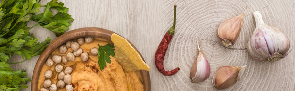vista superior del delicioso hummus con garbanzos, nacho en un tazón cerca de especias y perejil en una mesa de madera beige, plano panorámico
 - Foto, Imagen