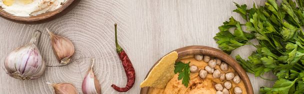 vista superior del delicioso hummus con garbanzos, nacho en un tazón cerca de especias y perejil en una mesa de madera beige, plano panorámico
 - Foto, Imagen