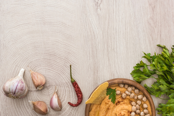 сверху вид на вкусный хумус с горохом, начо в миске рядом со специями и петрушку на бежевом деревянном столе
 - Фото, изображение