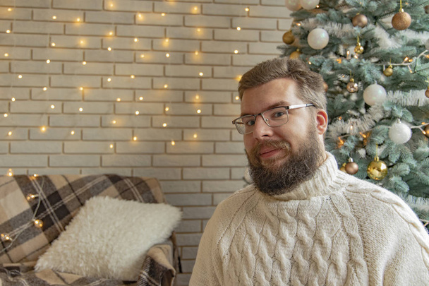 απλό πορτρέτο του μέσου ενήλικα γενειοφόρος άνθρωπος σε γυαλιά σε χριστουγεννιάτικο εσωτερικό διακοσμημένο περιβάλλον τούβλο τοίχο γιρλάντα κίτρινο φωτισμό φόντο, αντίγραφο χώρου  - Φωτογραφία, εικόνα