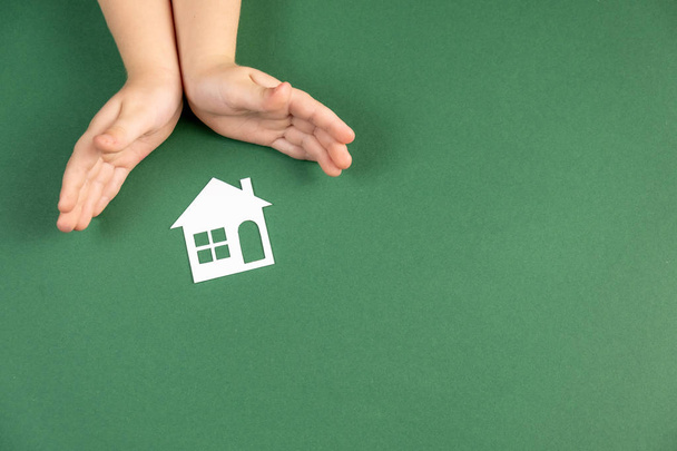 Dakloze opvang en onroerend goed concept. Close-up van kinderhanden met wit papieren huis en hart op groene achtergrond. Vlakke lay, kopieerruimte - Foto, afbeelding