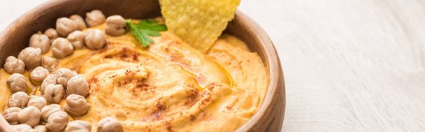 close up view of delicious hummus with chickpeas and nacho in bowl on beige wooden table, panoramic shot - Foto, Bild