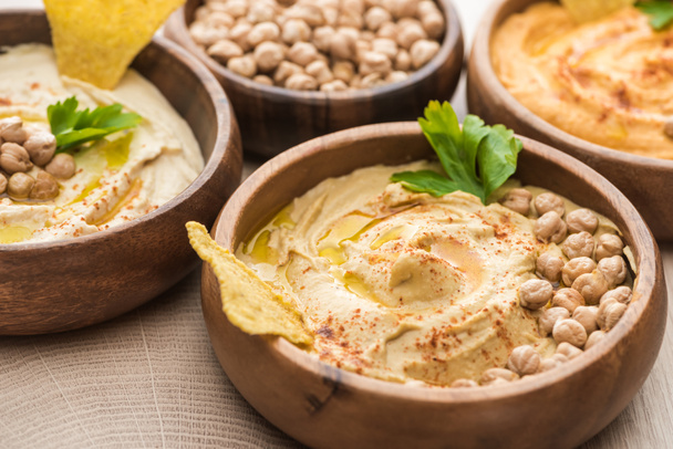 Nahaufnahme von köstlichem Hummus mit Kichererbsen und Nacho in Schalen auf beigem Holztisch - Foto, Bild