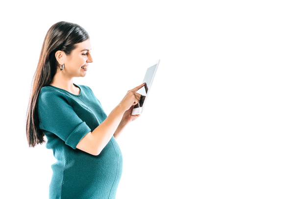 вид сбоку улыбающейся беременной девушки с помощью цифрового планшета, изолированного на белом
 - Фото, изображение