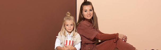 panoramic shot of daughter with popcorn and smiling mother looking at camera on beige and brown background  - Photo, Image
