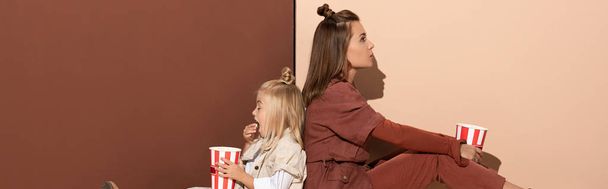 Panoramaaufnahme der Tochter beim Popcorn essen und der Mutter auf beigem und braunem Hintergrund  - Foto, Bild