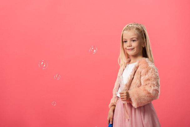 lindo y sonriente niño sosteniendo botella con burbujas de jabón aislado en rosa
 - Foto, Imagen