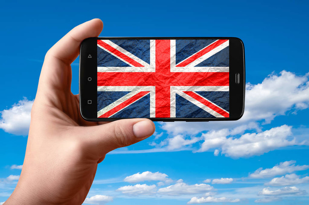 Bandera británica en la pantalla del teléfono. Smartphone en la mano muestra una bandera en un fondo del cielo con nubes
.  - Foto, imagen