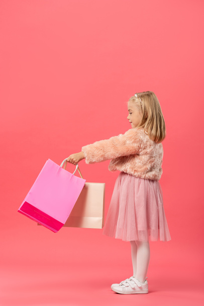 side view of cute and smiling kid holding shopping bags on pink background  - Valokuva, kuva
