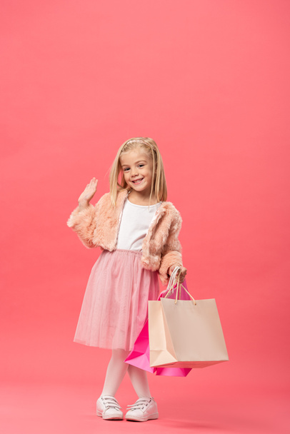 χαριτωμένο και χαμογελαστό παιδί χαιρετώντας και κρατώντας τσάντες ψώνια σε ροζ φόντο  - Φωτογραφία, εικόνα
