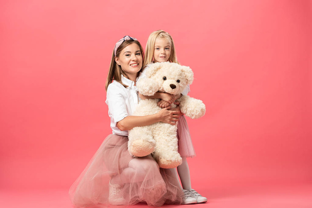 daughter and smiling mother with teddy bear on pink background  - Foto, Imagen
