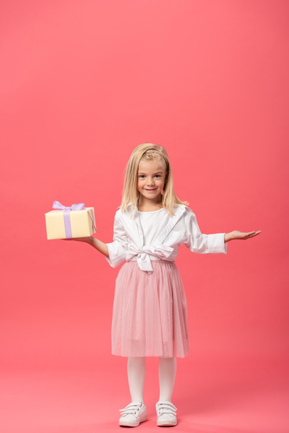 cute and smiling kid with outstretched hand holding gift on pink background  - Fotoğraf, Görsel