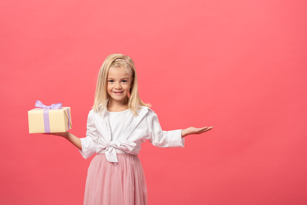 χαριτωμένο και χαμογελαστό παιδί με απλωμένο χέρι κρατώντας δώρο απομονώνονται σε ροζ - Φωτογραφία, εικόνα