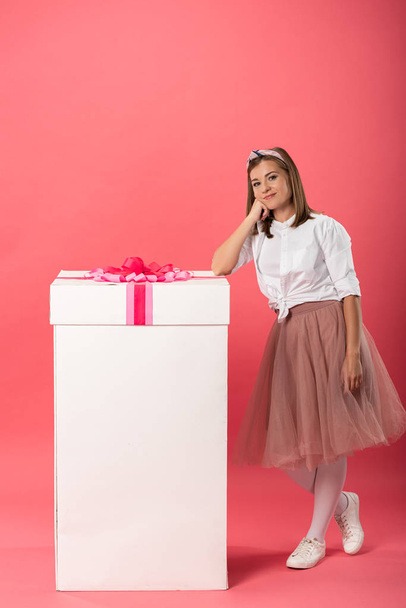 attractive and smiling woman standing near gift box on pink background  - Фото, изображение