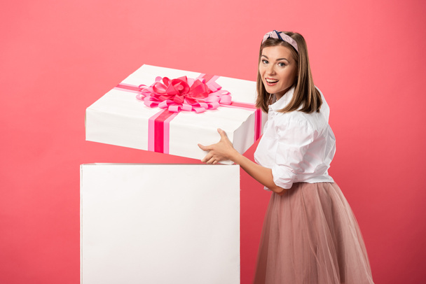 atrakcyjna i uśmiechnięta kobieta otwierając pudełko odizolowane na różowo - Zdjęcie, obraz