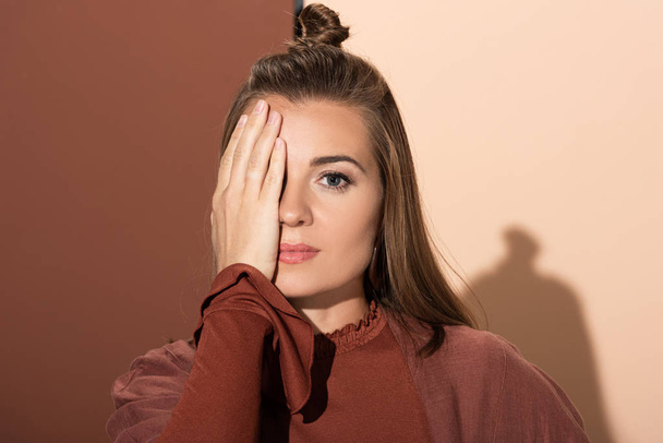 attractive woman obscuring face and looking at camera on beige and brown background  - Fotoğraf, Görsel