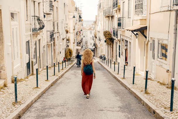 Egy fiatal szőke turista piros ruhában és kalapban felfedez egy belvárost egy európai városban. - Fotó, kép