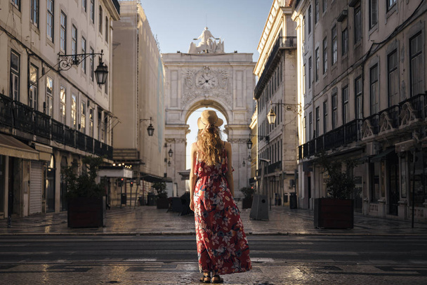 A young blond tourist in a red generic sundress and a hat is exploring a downtown on a European city - Фото, зображення