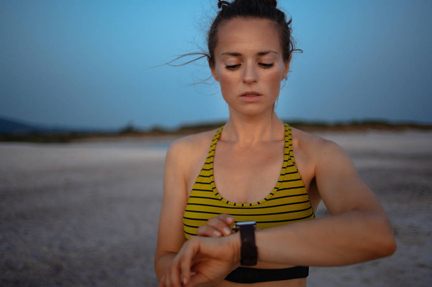 Portrait of fit sports woman in sport clothes on the seashore at sunset using smart watch to track steps, runs or rides in fitness app. - 写真・画像
