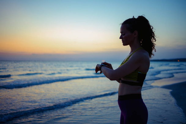Silhouette de femme sportive en bonne santé en vêtements de fitness sur la côte de l'océan le soir en utilisant la montre intelligente pour partager les résultats de l'entraînement dans les médias sociaux
. - Photo, image