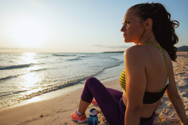 active sports woman in sport style clothes with bottle of water sitting and looking into the distance on the ocean shore in the evening. - Foto, afbeelding