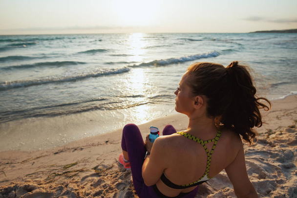 Seen from behind fitness woman in sport clothes with bottle of water relaxing after workout on the ocean coast in the evening. - Foto, Imagem