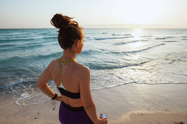 Seen from behind young sports woman in sport style clothes with bottle of water looking into the distance on the ocean coast at sunset. - Fotó, kép