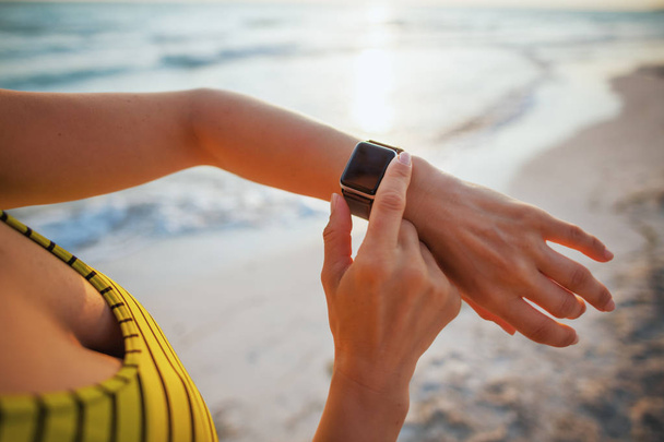 Closeup on young woman in sport style clothes on the beach at sunset using smart watch using app to track fitness activity. - Foto, Imagem