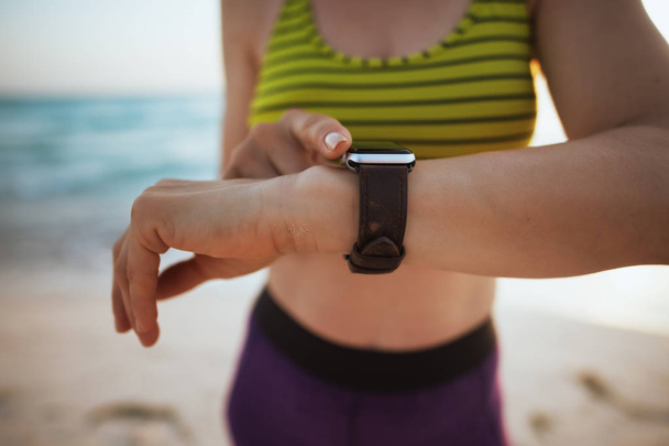 Primo piano sulla donna in forma in abiti sportivi sulla costa al tramonto con orologio intelligente utilizzando l'app per monitorare l'attività di fitness
. - Foto, immagini