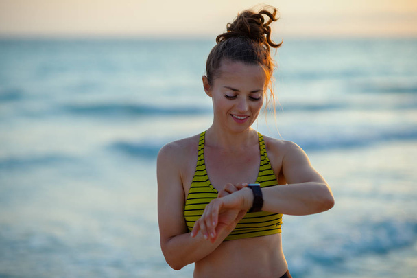 mulher esportiva ativa em roupas de estilo esportivo na praia ao pôr do sol usando relógio inteligente para rastrear passos, corridas ou passeios em aplicativo de fitness
. - Foto, Imagem