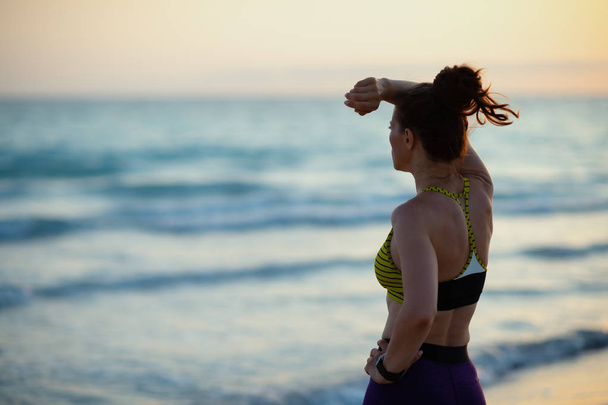 Seen from behind healthy sports woman in sport style clothes on the ocean coast at sunset looking into the distance. - Photo, image
