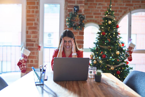 ストレスが原因で頭痛の種に手でクリスマスツリーの周りの自宅でノートパソコンで作業テーブルに座って美しい女性。片頭痛に苦しむ. - 写真・画像