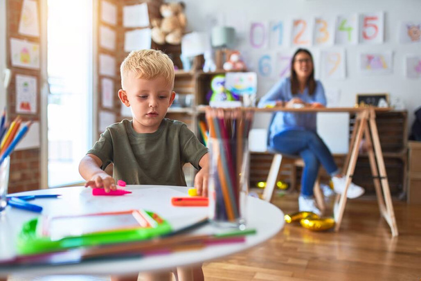 Young caucasian child playing at playschool with teacher. Mother and son at playroom drawing a draw with color pencils, young woman at the background sitting on desk. - Photo, Image
