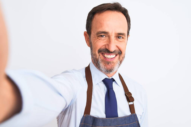 Middle age shopkeeper man wearing apron make selfie over isolated white background with a happy face standing and smiling with a confident smile showing teeth - Zdjęcie, obraz