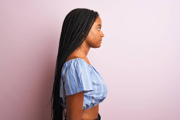Африканская американка в полосатой футболке стоя на изолированном розовом фоне глядя в сторону, расслабиться профиль позировать с естественным лицом с уверенной улыбкой
. - Фото, изображение