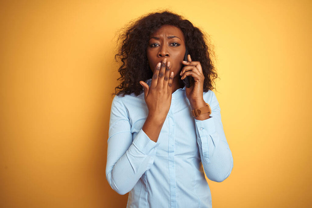 Young african american woman talking on the smartphone over isolated yellow background cover mouth with hand shocked with shame for mistake, expression of fear, scared in silence, secret concept - Foto, afbeelding