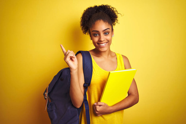American student woman wearing backpack holding notebook over isolated yellow background very happy pointing with hand and finger to the side - Photo, Image