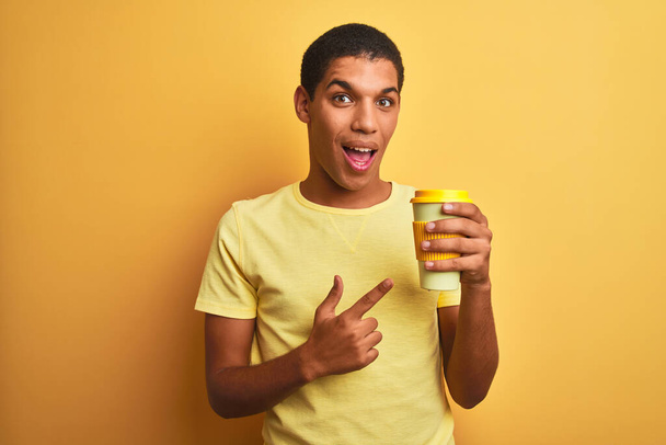 Genç, yakışıklı Arap adam izole edilmiş sarı arka planda kahve içerek elini ve parmağını göstererek mutlu oluyor. - Fotoğraf, Görsel